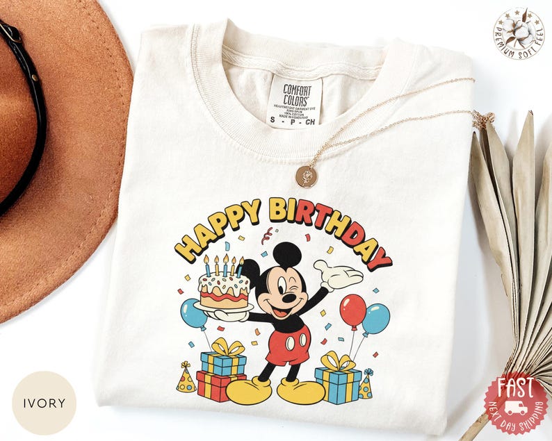 K&ouml;nnte beinhalten: Elfenbeinfarbenes T-Shirt mit einem farbenfrohen Mickey Mouse-Geburtstagsmotiv. Das Design zeigt einen Kuchen, Luftballons und den Schriftzug "HAPPY BIRTHDAY". Das T-Shirt hat einen Rundhalsausschnitt und kurze &Auml;rmel.