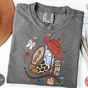 May include: A charcoal gray short-sleeve t-shirt featuring a graphic print of a cow-print cowboy boot, a red hat, and blocks with the letters T, S, and the number 13. The design includes a bow, flowers, and the name Taylor on the boot sole.