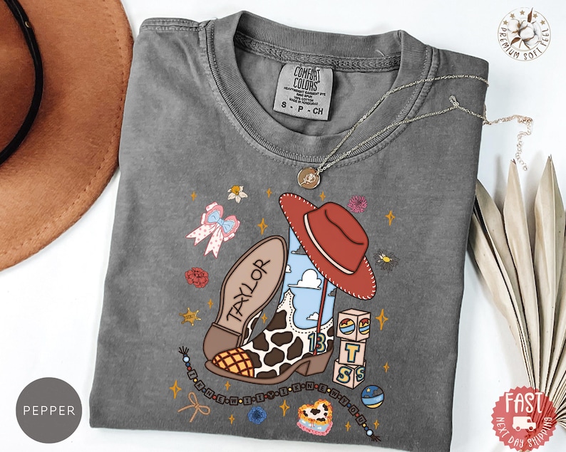 May include: A charcoal gray short-sleeve t-shirt featuring a graphic print of a cow-print cowboy boot, a red hat, and blocks with the letters T, S, and the number 13. The design includes a bow, flowers, and the name Taylor on the boot sole.