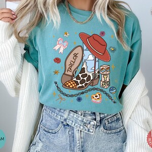 May include: A seafoam green t-shirt featuring a graphic print of a cowboy boot with a cow print pattern, a red cowboy hat, the name Taylor, the number 13, stacked blocks, a beaded necklace, a bow, and small floral accents in various colors.