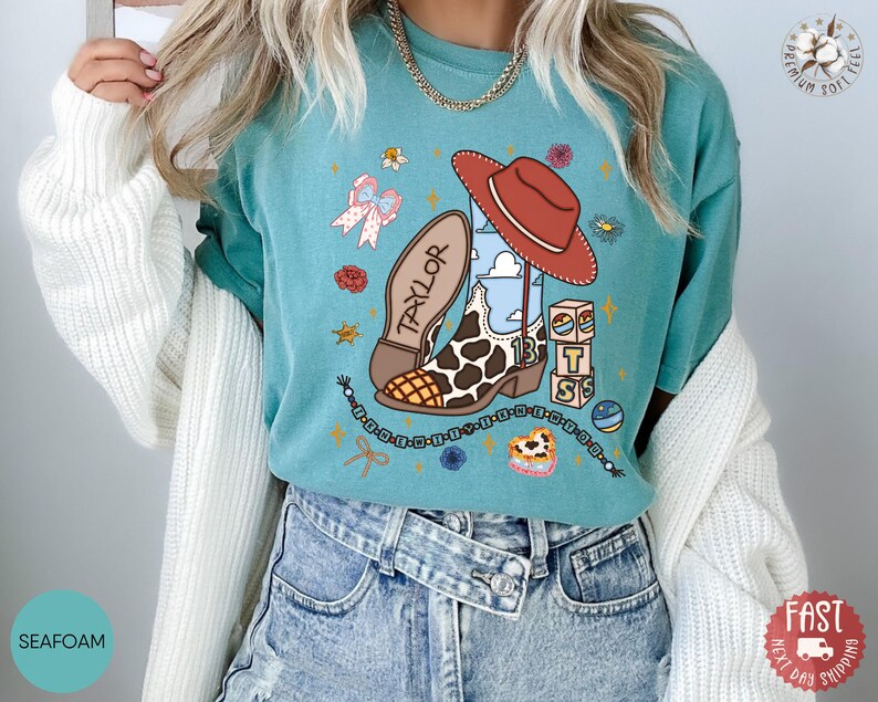 May include: A seafoam green t-shirt featuring a graphic print of a cowboy boot with a cow print pattern, a red cowboy hat, the name Taylor, the number 13, stacked blocks, a beaded necklace, a bow, and small floral accents in various colors.