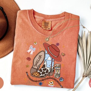 May include: A terracotta-colored crew neck t-shirt featuring a graphic print of a cow-print cowboy boot, a brown hat, a stack of blocks with the letters T, S, and a cloud design, a bow, and the name Taylor written on the boot sole.