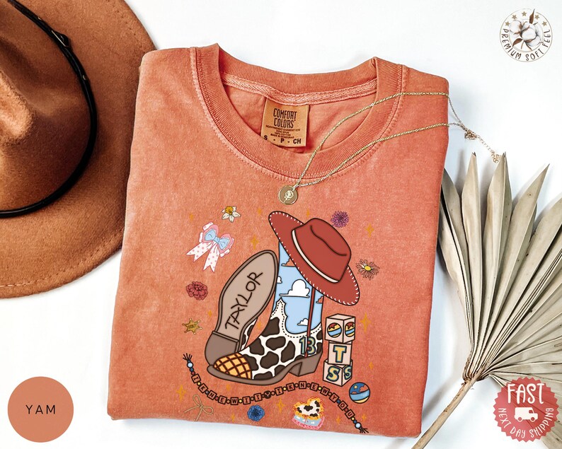 May include: A terracotta-colored crew neck t-shirt featuring a graphic print of a cow-print cowboy boot, a brown hat, a stack of blocks with the letters T, S, and a cloud design, a bow, and the name Taylor written on the boot sole.