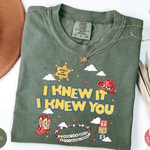 May include: A moss green t-shirt featuring a graphic print with the text I KNEW IT I KNEW YOU, a yellow sheriff star with the text INFINITY AND BEYOND, a red cowboy hat, a pair of red cowboy boots, a beaded bracelet, and a stack of lettered blocks.