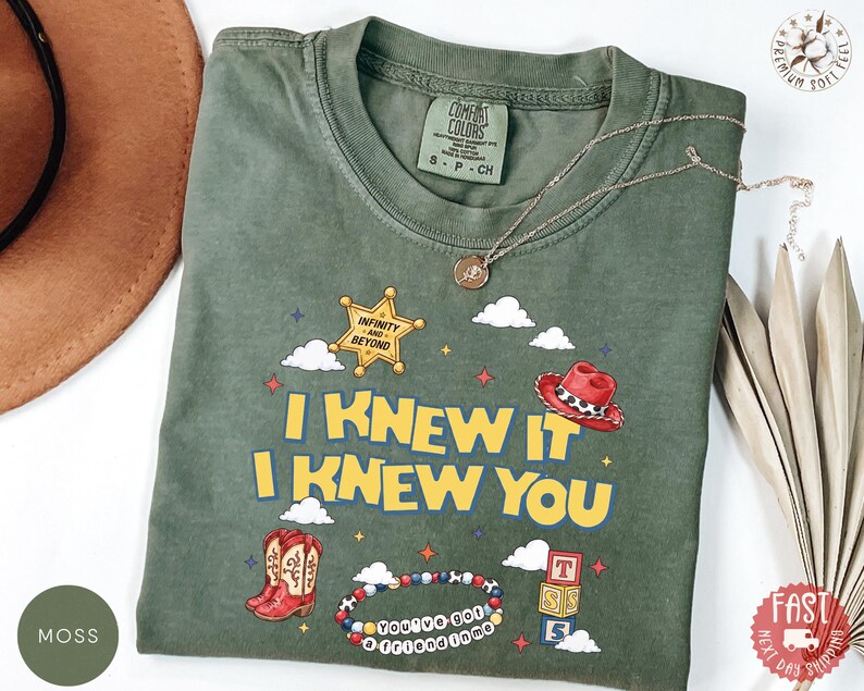 May include: A moss green t-shirt featuring a graphic print with the text I KNEW IT I KNEW YOU, a yellow sheriff star with the text INFINITY AND BEYOND, a red cowboy hat, a pair of red cowboy boots, a beaded bracelet, and a stack of lettered blocks.