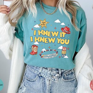 May include: A seafoam green t-shirt featuring a graphic print with the text I KNEW IT I KNEW YOU. The design includes a yellow sheriff star, a red cowboy hat, red cowboy boots, a beaded bracelet with text, and stacked alphabet blocks.