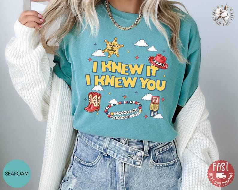 May include: A seafoam green t-shirt featuring a graphic print with the text I KNEW IT I KNEW YOU. The design includes a yellow sheriff star, a red cowboy hat, red cowboy boots, a beaded bracelet with text, and stacked alphabet blocks.