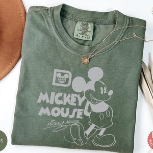 May include: Moss green t-shirt with a Mickey Mouse graphic and the words "MICKEY MOUSE". The shirt has a crew neck and short sleeves. A gold necklace with a pendant is draped across the shirt. The shirt is made of a soft material.