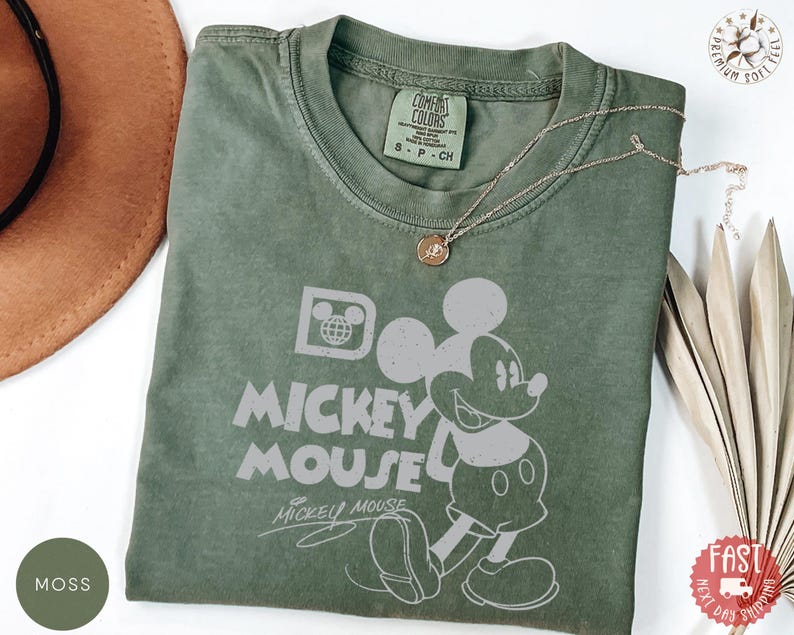 May include: Moss green t-shirt with a Mickey Mouse graphic and the words "MICKEY MOUSE". The shirt has a crew neck and short sleeves. A gold necklace with a pendant is draped across the shirt. The shirt is made of a soft material.