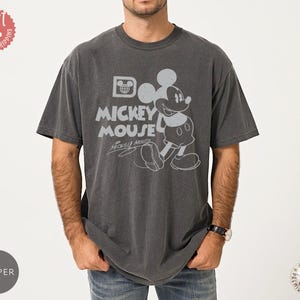 May include: Dark grey t-shirt with a distressed Mickey Mouse graphic. The design includes the words "Mickey Mouse" and Mickey's signature. The shirt has a vintage, worn appearance.