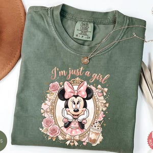 May include: A moss green short-sleeve t-shirt featuring a graphic of Minnie Mouse inside an ornate gold frame, surrounded by pink roses and bows. The text "I'm just a girl" is printed above the frame in a cursive font, with a small iced coffee illustration.