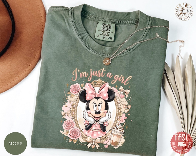 May include: A moss green short-sleeve t-shirt featuring a graphic of Minnie Mouse inside an ornate gold frame, surrounded by pink roses and bows. The text "I'm just a girl" is printed above the frame in a cursive font, with a small iced coffee illustration.