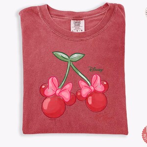 Peut inclure: T-shirt rouge avec un motif de cerises orn&eacute; de n&oelig;uds de Minnie Mouse et du logo Disney. Le t-shirt a un col rond et est fait d'une mati&egrave;re douce. L'&eacute;tiquette Comfort Colors est visible en haut.