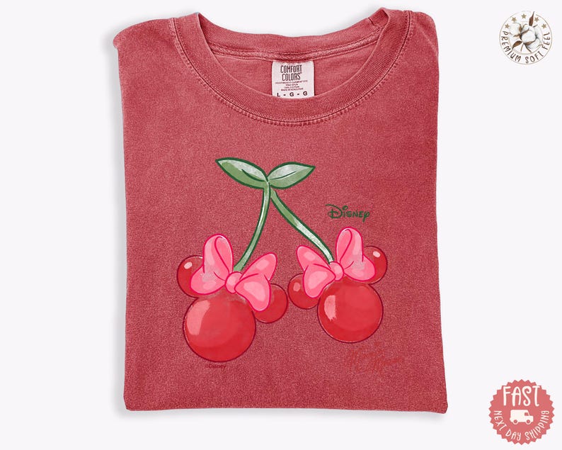 Peut inclure: T-shirt rouge avec un motif de cerises orn&eacute; de n&oelig;uds de Minnie Mouse et du logo Disney. Le t-shirt a un col rond et est fait d'une mati&egrave;re douce. L'&eacute;tiquette Comfort Colors est visible en haut.