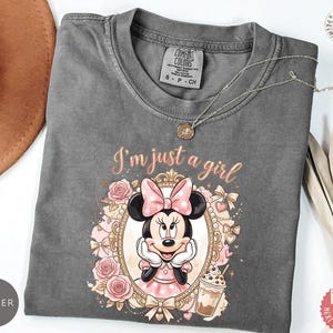 May include: A charcoal gray t-shirt featuring a graphic of Minnie Mouse inside an ornate gold frame, surrounded by pink roses and bows. The text "I'm just a girl" is printed above the frame in a cursive font. A small iced coffee drink is visible.