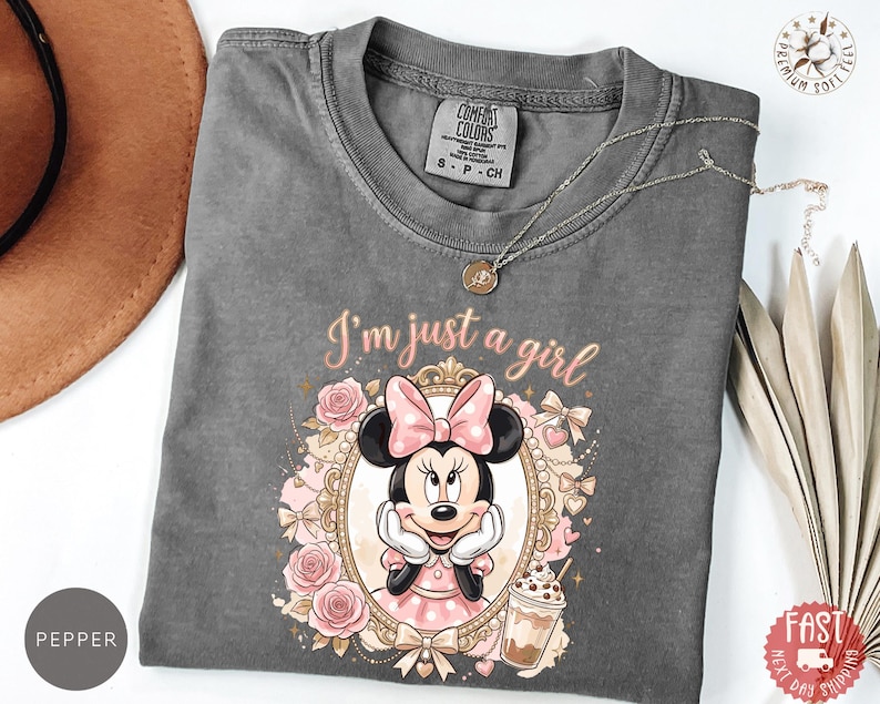 May include: A charcoal gray t-shirt featuring a graphic of Minnie Mouse inside an ornate gold frame, surrounded by pink roses and bows. The text "I'm just a girl" is printed above the frame in a cursive font. A small iced coffee drink is visible.