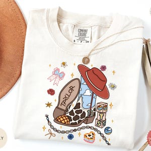 May include: An ivory t-shirt featuring a graphic print of a cow-print cowboy boot with the name Taylor, a red cowboy hat, building blocks with the letters BTS, a beaded necklace, a pink bow, flowers, and stars. The shirt has a Comfort Colors label.