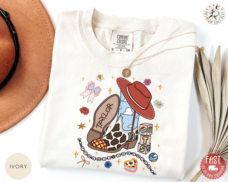 May include: An ivory t-shirt featuring a graphic print of a cow-print cowboy boot with the name Taylor, a red cowboy hat, building blocks with the letters BTS, a beaded necklace, a pink bow, flowers, and stars. The shirt has a Comfort Colors label.