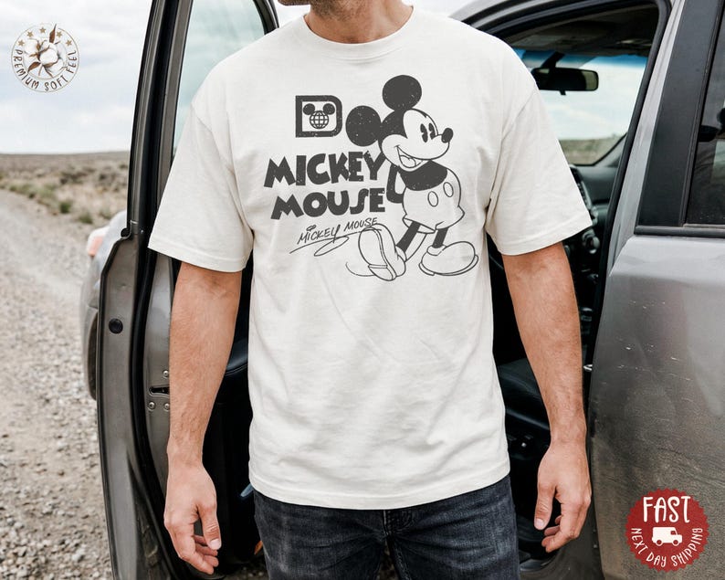 May include: Cream-coloured t-shirt with a vintage Mickey Mouse graphic. The design includes Mickey Mouse's image, the words "Mickey Mouse," and his signature in black. The shirt is short-sleeved and appears to be made of soft material.