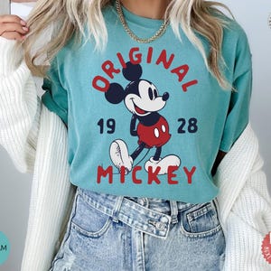 May include: Seafoam green t-shirt featuring a graphic of Mickey Mouse with the words "ORIGINAL MICKEY" in red. The design includes the numbers "19" and "28". The shirt is made of soft material.