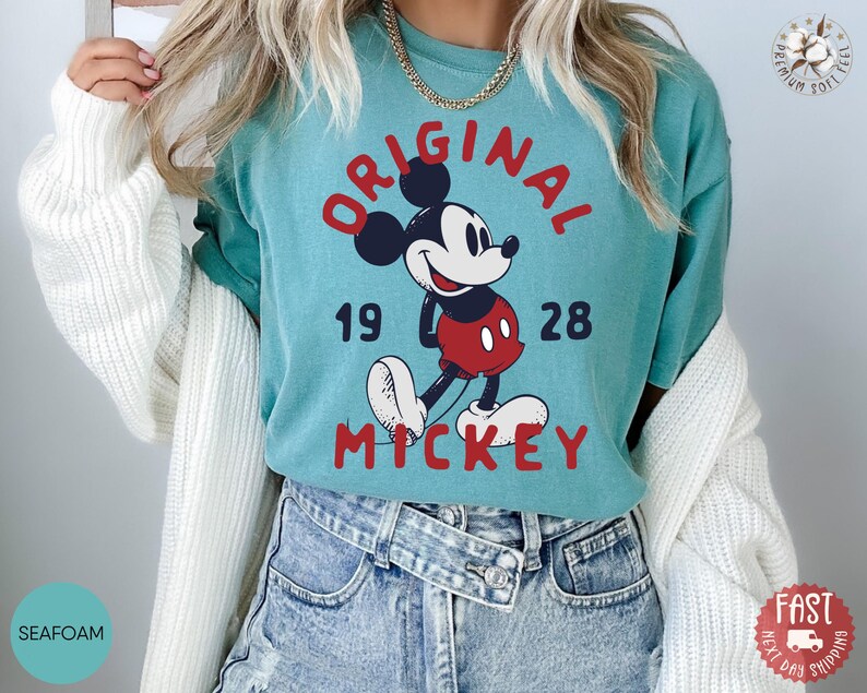 May include: Seafoam green t-shirt featuring a graphic of Mickey Mouse with the words "ORIGINAL MICKEY" in red. The design includes the numbers "19" and "28". The shirt is made of soft material.