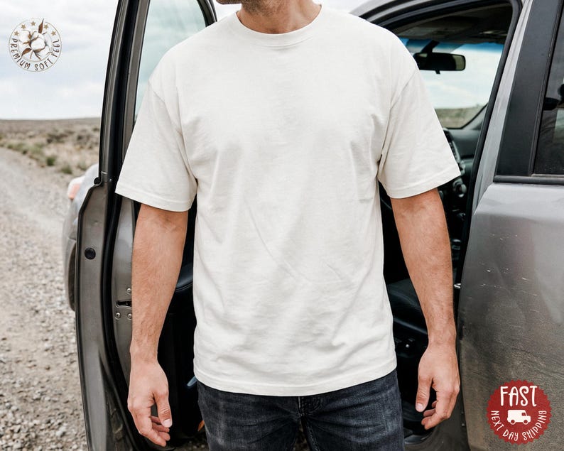 May include: A solid off-white short-sleeve t-shirt with a crew neckline. The shirt features a plain, unpatterned design with a standard fit, short sleeves, and a straight hemline, worn over dark gray denim jeans in a casual outdoor setting.