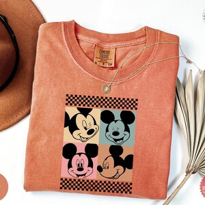 Peut inclure: T-shirt corail Comfort Colors avec un motif pop art de Mickey Mouse en quatre expressions diff&eacute;rentes, encadr&eacute; d'un motif &agrave; carreaux noir et blanc. Un collier dor&eacute; est pos&eacute; sur le t-shirt. Le t-shirt a une &eacute;tiquette Comfort Colors.
