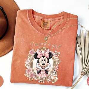 May include: A terracotta-colored crew neck t-shirt featuring a graphic of Minnie Mouse inside an ornate gold frame, surrounded by pink roses and bows. The text "I'm just a girl" is printed above the frame, accompanied by a small iced coffee illustration.