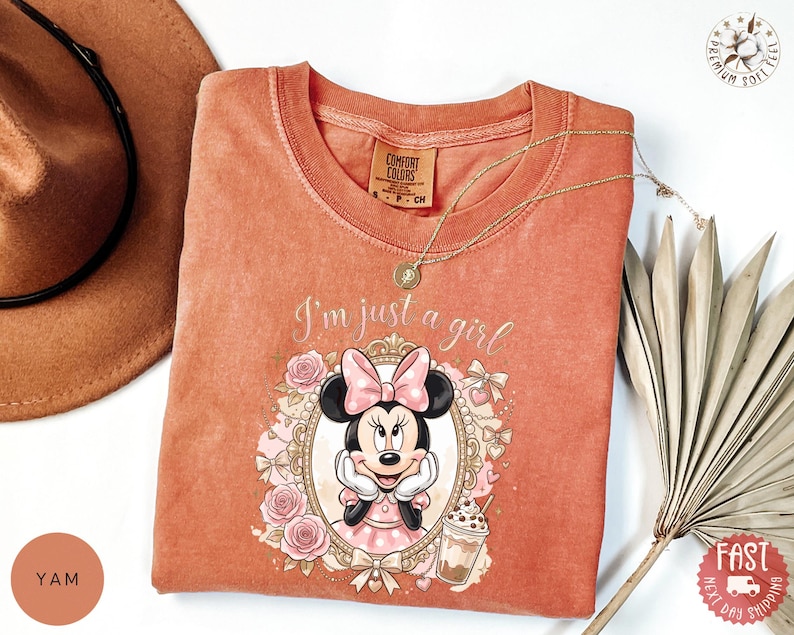 May include: A terracotta-colored crew neck t-shirt featuring a graphic of Minnie Mouse inside an ornate gold frame, surrounded by pink roses and bows. The text "I'm just a girl" is printed above the frame, accompanied by a small iced coffee illustration.