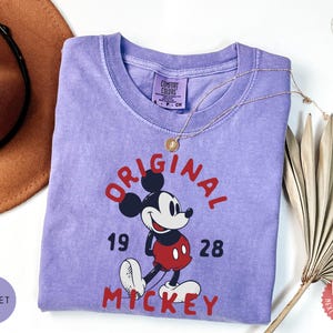 May include: A violet Comfort Colors t-shirt featuring a graphic of Mickey Mouse with the words "ORIGINAL MICKEY 1928" in red. A gold necklace and a brown hat are next to the shirt. The shirt has a crew neck and short sleeves.