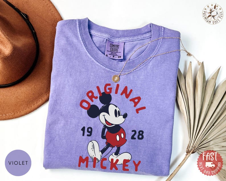 May include: A violet Comfort Colors t-shirt featuring a graphic of Mickey Mouse with the words "ORIGINAL MICKEY 1928" in red. A gold necklace and a brown hat are next to the shirt. The shirt has a crew neck and short sleeves.
