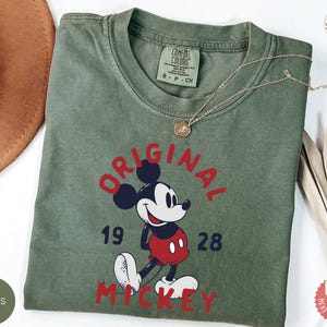 May include: Moss green t-shirt featuring a graphic of Mickey Mouse with the text "ORIGINAL MICKEY 1928" in red. The shirt has a crew neck and short sleeves. A gold necklace is draped across the shirt.