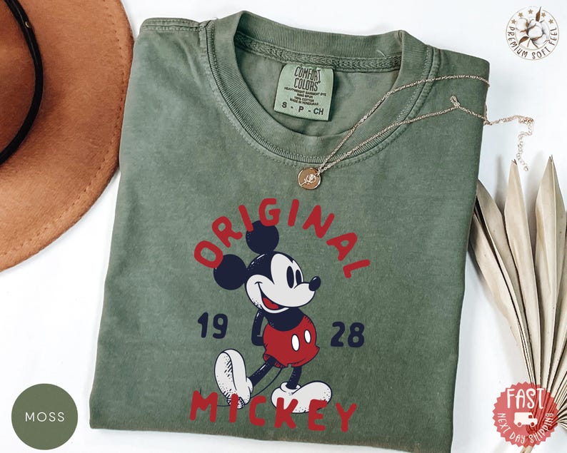 May include: Moss green t-shirt featuring a graphic of Mickey Mouse with the text "ORIGINAL MICKEY 1928" in red. The shirt has a crew neck and short sleeves. A gold necklace is draped across the shirt.
