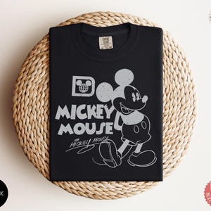 May include: Black t-shirt with a distressed Mickey Mouse graphic and the words "Mickey Mouse". The design includes Mickey's image and the Disney logo. The shirt is displayed on a woven, circular surface.