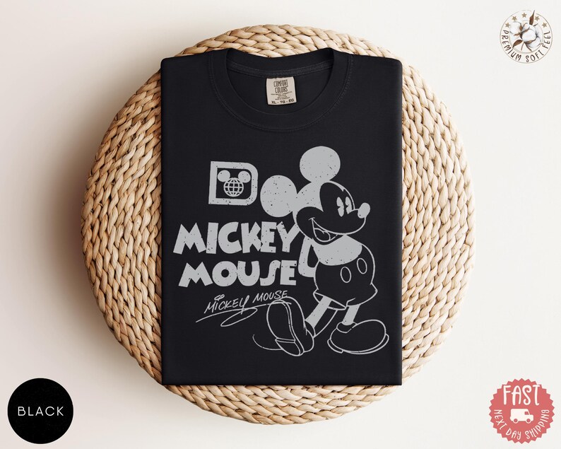 May include: Black t-shirt with a distressed Mickey Mouse graphic and the words "Mickey Mouse". The design includes Mickey's image and the Disney logo. The shirt is displayed on a woven, circular surface.