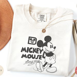 May include: Cream-coloured t-shirt with a black and white Mickey Mouse graphic. The design includes the words "Do Mickey Mouse" and Mickey's signature. A gold necklace and a brown hat are also visible.