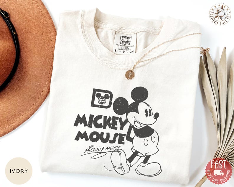 May include: Cream-coloured t-shirt with a black and white Mickey Mouse graphic. The design includes the words "Do Mickey Mouse" and Mickey's signature. A gold necklace and a brown hat are also visible.