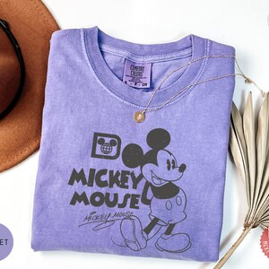 May include: A violet Comfort Colors t-shirt with a black Mickey Mouse graphic and the text "Do Mickey Mouse". A gold necklace is on the shirt. A brown hat and dried leaves are in the background. The shirt is a Comfort Colors brand.