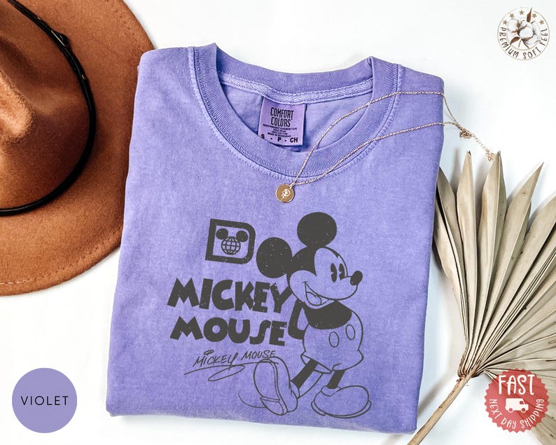 May include: A violet Comfort Colors t-shirt with a black Mickey Mouse graphic and the text "Do Mickey Mouse". A gold necklace is on the shirt. A brown hat and dried leaves are in the background. The shirt is a Comfort Colors brand.