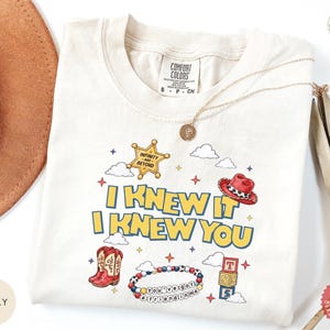 May include: An ivory t-shirt featuring a graphic print with the text I KNEW IT I KNEW YOU. The design includes a yellow sheriff star badge, a red cowboy hat, red cowboy boots, a beaded bracelet with text, alphabet blocks, and clouds.