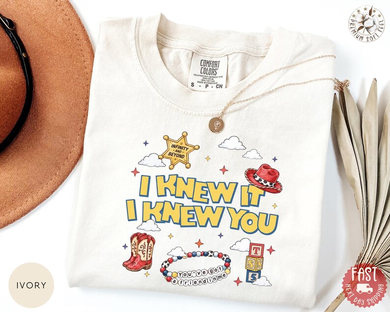 May include: An ivory t-shirt featuring a graphic print with the text I KNEW IT I KNEW YOU. The design includes a yellow sheriff star badge, a red cowboy hat, red cowboy boots, a beaded bracelet with text, alphabet blocks, and clouds.