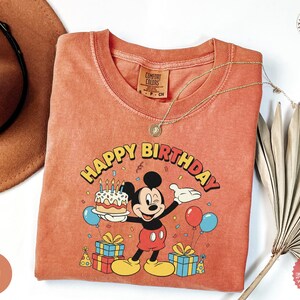 K&ouml;nnte beinhalten: Orange-rotes T-Shirt mit einer Mickey Mouse-Grafik, die eine Geburtstagstorte h&auml;lt, mit den Worten "Happy Birthday". Das T-Shirt hat einen Rundhalsausschnitt und kurze &Auml;rmel. Eine goldene Halskette ist &uuml;ber dem T-Shirt drapiert. Das T-Shirt ist auf einer wei&szlig;en Oberfl&auml;che gefaltet.