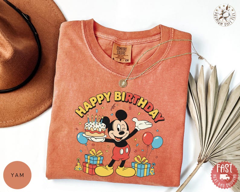 K&ouml;nnte beinhalten: Orange-rotes T-Shirt mit einer Mickey Mouse-Grafik, die eine Geburtstagstorte h&auml;lt, mit den Worten "Happy Birthday". Das T-Shirt hat einen Rundhalsausschnitt und kurze &Auml;rmel. Eine goldene Halskette ist &uuml;ber dem T-Shirt drapiert. Das T-Shirt ist auf einer wei&szlig;en Oberfl&auml;che gefaltet.