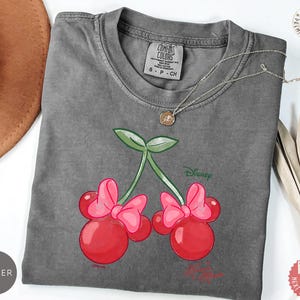 Peut inclure: Un t-shirt gris Comfort Colors avec un motif de cerises avec des n&oelig;uds roses et des feuilles vertes. Le design comprend le mot "Disney". Un collier argent&eacute; est drap&eacute; sur le t-shirt.