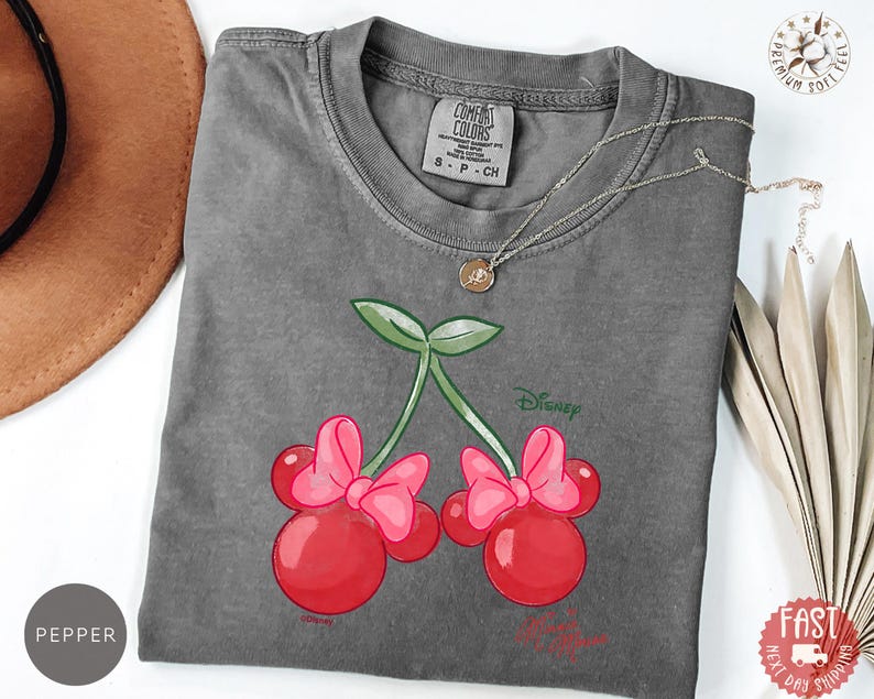 Peut inclure: Un t-shirt gris Comfort Colors avec un motif de cerises avec des n&oelig;uds roses et des feuilles vertes. Le design comprend le mot "Disney". Un collier argent&eacute; est drap&eacute; sur le t-shirt.