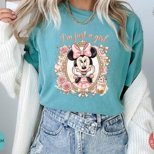 May include: A seafoam green t-shirt featuring a graphic of Minnie Mouse inside an ornate gold frame, surrounded by pink roses and bows. The text "I'm just a girl" is printed in a cursive font above the character, who is holding a coffee cup.