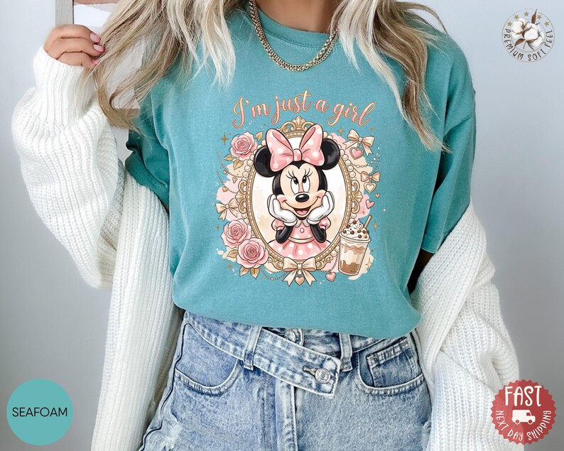 May include: A seafoam green t-shirt featuring a graphic of Minnie Mouse inside an ornate gold frame, surrounded by pink roses and bows. The text "I'm just a girl" is printed in a cursive font above the character, who is holding a coffee cup.