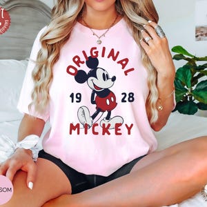 May include: A light pink t-shirt featuring a graphic of Mickey Mouse with the text "ORIGINAL MICKEY 1928" in red. The shirt has a crew neck and short sleeves. The image also includes a "FAST NEXT DAY SHIPPING" sticker.