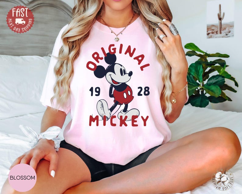 May include: A light pink t-shirt featuring a graphic of Mickey Mouse with the text "ORIGINAL MICKEY 1928" in red. The shirt has a crew neck and short sleeves. The image also includes a "FAST NEXT DAY SHIPPING" sticker.