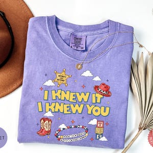 May include: A violet short-sleeved t-shirt featuring a graphic print with the text I KNEW IT I KNEW YOU, a yellow sheriff star, a red cowboy hat, red cowboy boots, a beaded bracelet with the text YOU'VE GOT A FRIEND IN ME, and alphabet blocks.